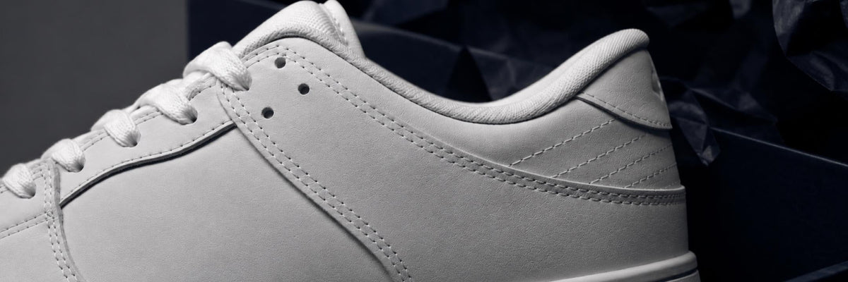 A close-up of a light gray leather sneaker, showing the side panel, white laces, and heel against a dark background.