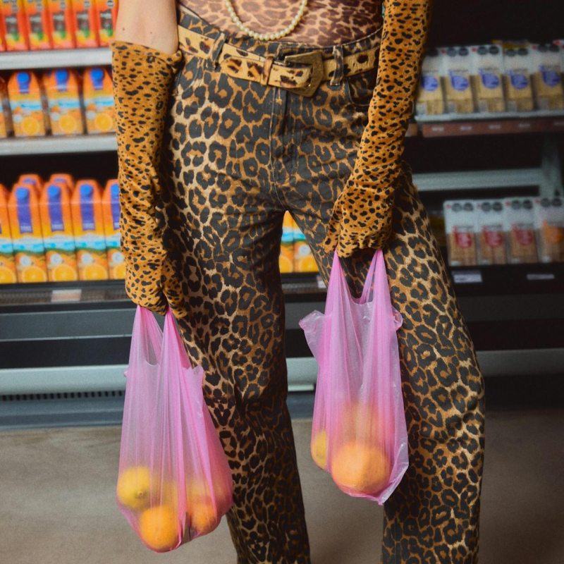 Person wearing leopard print pants holding pink bags with oranges in a grocery store.